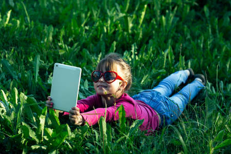 Little funny girl makes a selfie on a tablet in the green grass.の写真素材