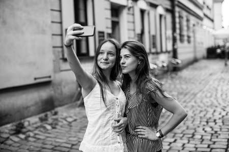 Two cute teenage girls take selfie on the street of city old district. Black and white photo.