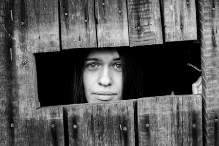 Young woman looking through the crack of a locked wooden shed, close-up. Black and white photo.の素材 [FY310170156935]