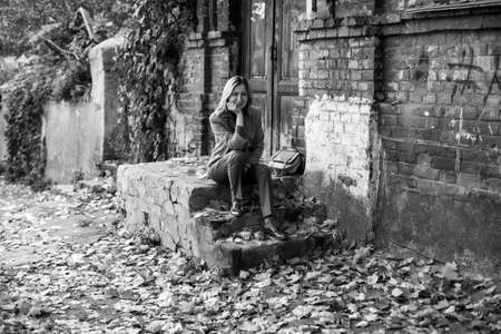 Elegant woman in a jacket sits on the porch of an old house with maple leaves on the ground in the autumn day atmosphere. Black and white photo.の写真素材