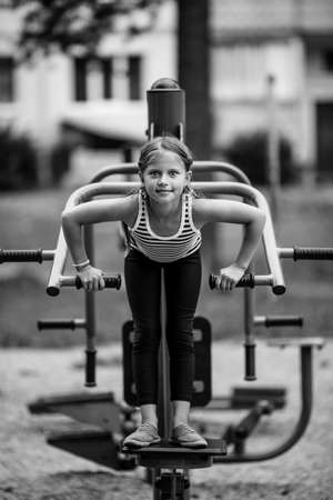 Little girl fooling on a trainer-machine on the playground outdoors. Black and white photo.の写真素材