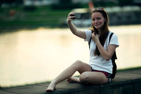 A teen girl takes a selfie while sitting on the river embankment.の写真素材