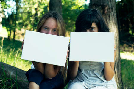 Two teen girl holding clean white sheet paper (banner for your message) in the park.の写真素材