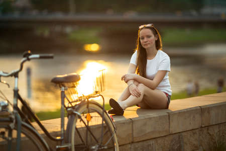 Cute girl with the bike sitting on the river promenade at amazing sunset.の写真素材