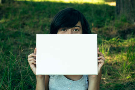 Teen girl holding clean white sheet paper (banner for your message), in the park at summer.の写真素材