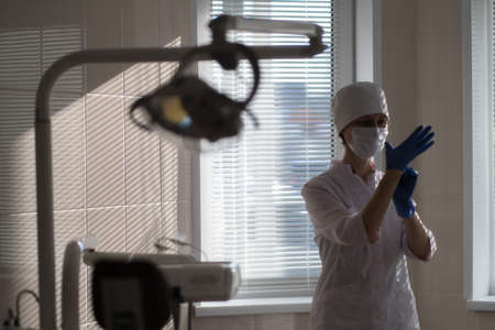 A female dentist in her medical office wears blue uniform gloves on her hands. In the soft natural...