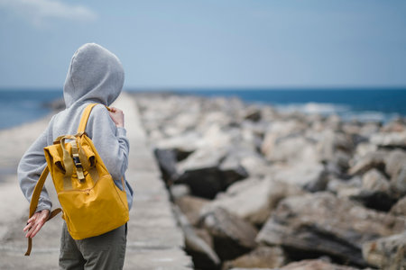 A hooded woman puts on a backpack while standing on an ocean pier. back view.