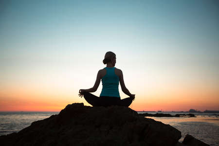 Woman practicing yoga in lotus pose on the coast during sunset.の写真素材