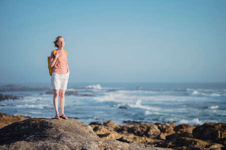 A woman with a backpack stands on the rocks near the sea.の写真素材