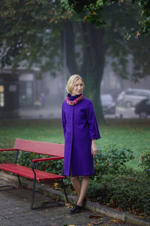 A woman in a lilac coat stands near a park bench.の写真素材