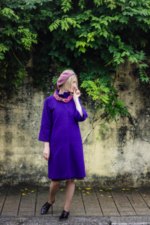A woman in a coat and beret poses by a wall covered with greenery.の写真素材