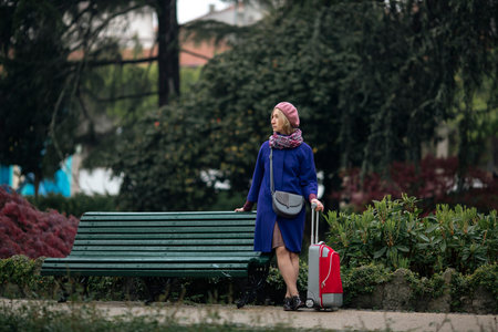A woman is standing near a bench in the park. She is carrying a red suitcase.の写真素材