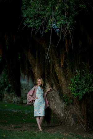 A woman in a summer dress stands near a tree in a city park.の写真素材