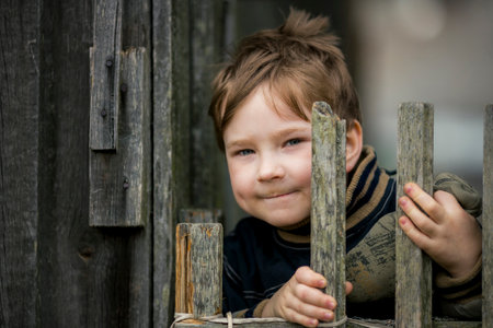 A small country boy peeks out from behind a wooden fence.の写真素材