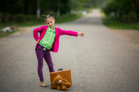 A teenage girl in bright clothes standing on the road with a suitcase and a stuffed toy, raising her thumb.の写真素材