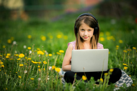 A teenage girl in headphones sits on a lawn with a laptop in a village, blending technology with...