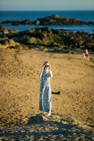 A full-length portrait of a woman in a summer dress and sunglasses standing on the rocks of an ocean shoreline, looking at the camera.の写真素材