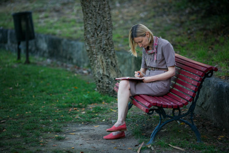 A woman sits on a park bench writing in a notebook or journal with a pen.の写真素材