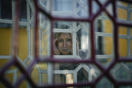 A woman gazes through a window with antique grilles in an old estate.の写真素材