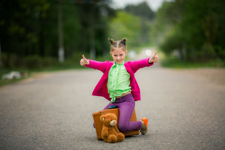 A little girl sits on a suitcase, giving a thumbs-up with a soft toy by her side.の写真素材
