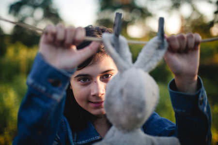 A teenage girl in a denim jacket stands outdoors, watching an old bunny toy hanging from a...