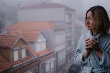 A woman in a bathrobe stands on her balcony, sipping tea as she gazes out at the street, shrouded in thick morning fog, creating a peaceful, dreamlike scene.の写真素材