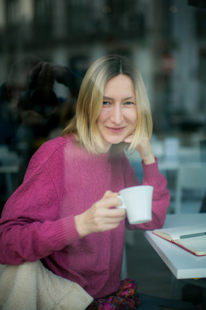 A portrait of a woman sitting in a cafÃ©, holding a cup and a notebook, while a photographer's reflection is visible in the window glass.の写真素材