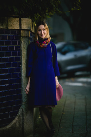 A woman in a coat, neck scarf stands near a wall, holding her beret in her hand, on an autumn city street.の写真素材
