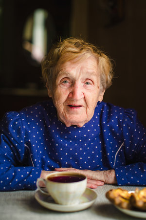 An elderly woman drinks tea at home with some pastries, enjoying a quiet moment in her cozy kitchen.の写真素材