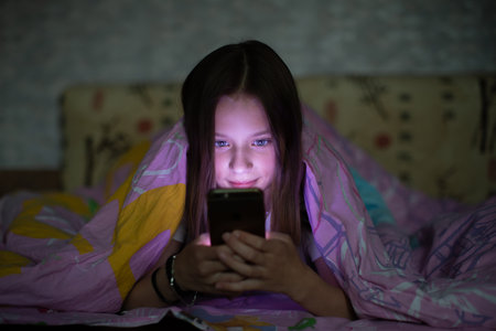 A teenage girl lying under a blanket, browsing her smartphone, immersed in the glow of the screen.の写真素材