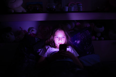 A 10-11-year-old girl browsing her smartphone under the blanket at night, illuminated by the screen's glow.の写真素材