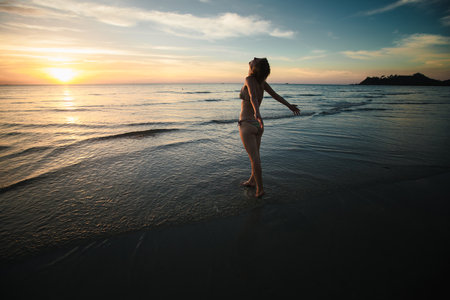 A woman in a swimsuit greets the sunrise on the beach, embodying relaxation and tranquility in a serene morning setting.の写真素材