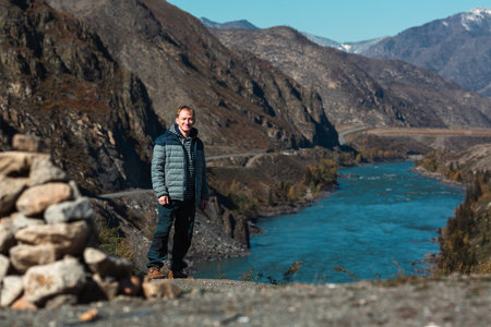 A man stands by a river in the mountains of the Altai Republic, Russia, surrounded by a rugged landscape.の写真素材