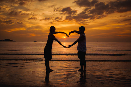 The silhouette of a couple on a seaside beach at twilight, forming a heart with their hands against the backdrop of the setting sun.の写真素材