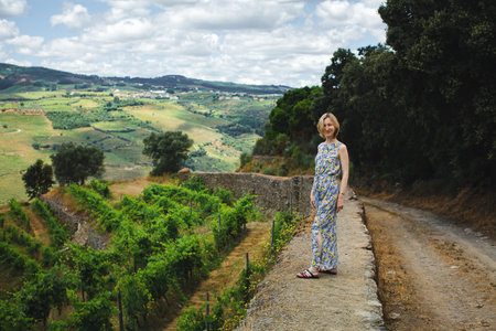 A woman in a summer dress walks along the stone wall of a vineyard in the Douro Valley, Portugal, surrounded by golden hills and timeless scenery.の写真素材