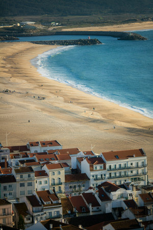 In the foreground, traditional Portuguese houses frame a sunlit beach in NazarÃ©, Portugal, on a clear day, where golden sands meet the serene Atlantic Ocean under a bright blue sky.の写真素材