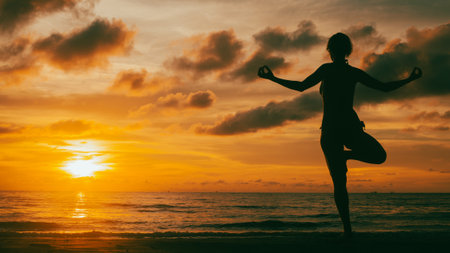 The silhouette of a woman practicing yoga on the beach during a breathtaking sunset. Her pose is steady and graceful, framed by glowing skies and the rhythm of the sea.の写真素材
