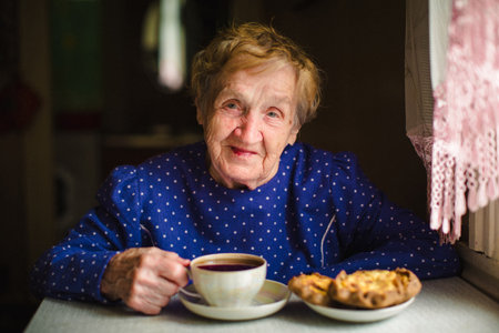 An elderly woman peacefully drinks tea in her home, her face calm and content. The portrait captures a gentle, warm moment filled with comfort, simplicity, and quiet joy.の写真素材