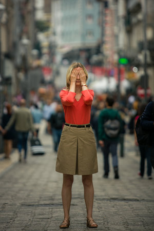 A woman stands frozen in the middle of a crowded street, covering her eyes tightly with her palms. Surrounded by the rush of people, she seems overwhelmed, trapped in a sudden panic as the world spins around her.の写真素材