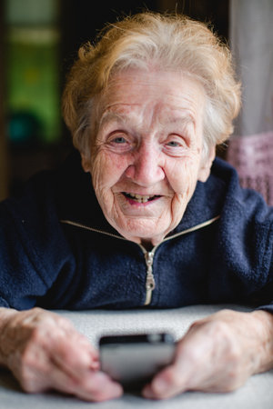 A joyful portrait of an elderly woman holding a smartphone in her hands. Her bright smile reflects...