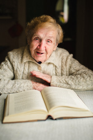 An elderly grandmother sits at a table at home reading a thick book, her face calm and focused. The warm scene reflects wisdom, patience, and the quiet joy of learning.の写真素材
