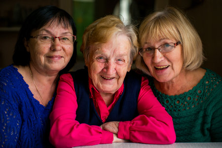 Warm portrait of three elderly women sitting close together, smiling at the camera, showing friendship, family bonds, and shared joy in a cozy indoor setting.の写真素材
