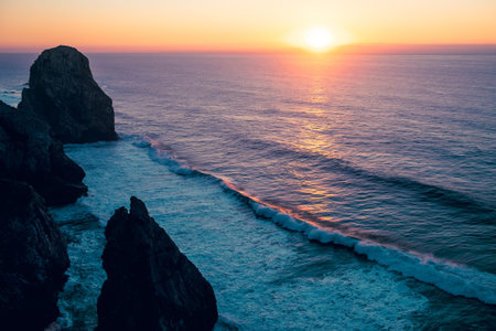 Sunset over Atlantic Ocean with waves rolling towards dark coastal cliffs, warm orange light...