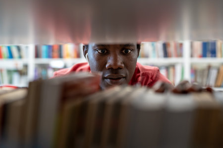 Black student man choosing textbook while standing between bookshelves in university libraryの写真素材