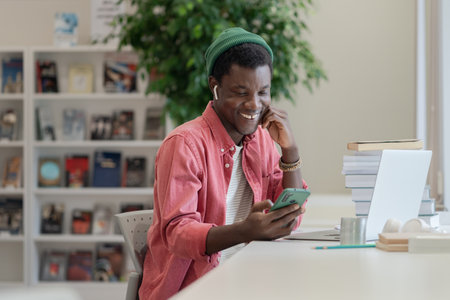 Happy African American student guy using mobile device while studying in libraryの写真素材