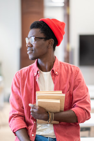 Young happy African American man university teacher sitting in classroom holding pile of booksの写真素材