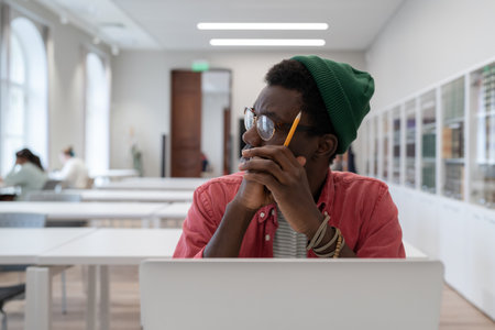 African student guy distracted from study, sitting at table with laptop in library and looking aside