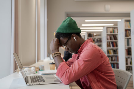 Tired apathetic African American man sits at table in library with laptop with eyes closedの写真素材