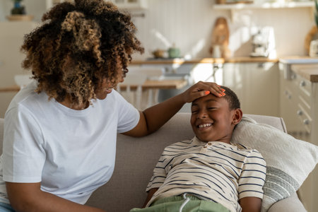 Caring kind African American mother strokes head praises smiling contented son boy sitting on couchの写真素材