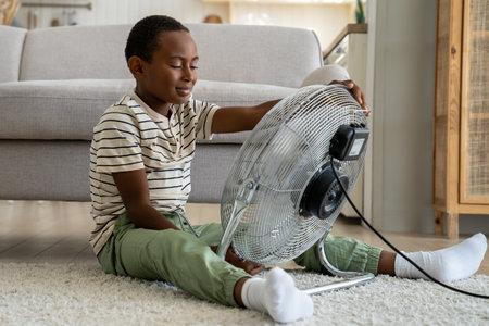 Calm African boy refreshing with electric fan at home alone. Small boy enjoy strong fresh wind blow